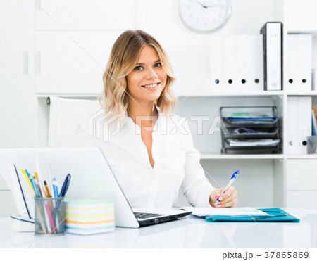 Young woman in clothes is sitting at the table in the office 37865869