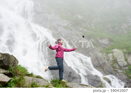 Smiling young female making selfie posing on rock in front of magnificent Balea waterfall in Romania 39041007