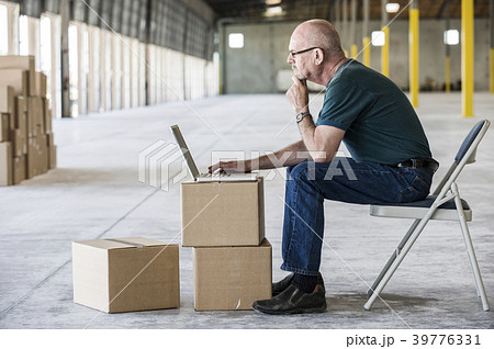 Caucasian man using boxes as desk and working on lap top computer in front of loading dock doors in a new warehouse. 39776331