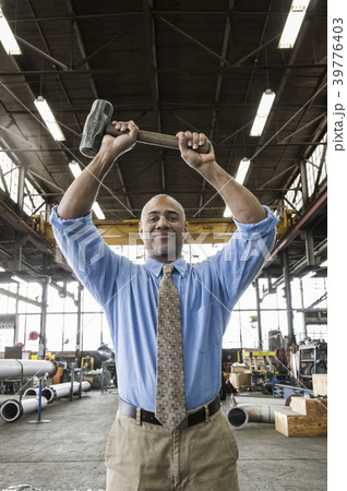 Black man owner of a sheet metal factory holding a hammer while standing on the floor of the factory. 39776403