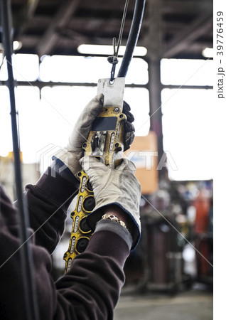 Factory worker using an overhead lift control unit to raise steel pipes in a sheet metal factory. 39776459