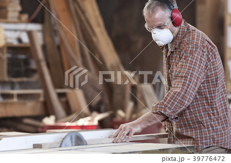 Caucasian man factory worker wearing hearing protection and a nose dust mask while cutting wood on a table saw. 39776472