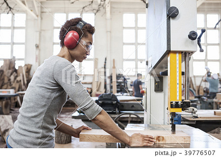 Black woman factory worker using a band saw to cut wood in a woodworking factory. Black woman factory worker using a band saw to cut wood in a woodworking factory. 39776507