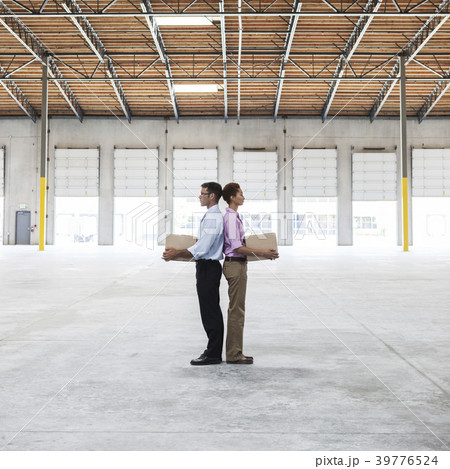 Caucasian man and black female holding cardboard boxes in middle of empty warehouse interior. 39776524