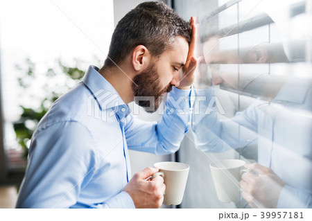 An unhappy man by the window holding a cup of coffee. 39957181