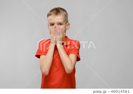 Portrait of blonde caucasian sad little boy in orange t-shirt closing his mouth with hands on gray Portrait of blonde caucasian sad little boy in orange t-shirt closing his mouth with hands on gray 40014561