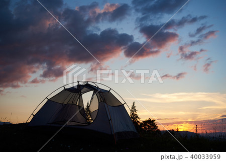 Camping at sunset in the mountains. The tent stands on grass with wildflowers under evening sky Camping at sunset in the mountains. The tent stands on grass with wildflowers under evening sky 40033959