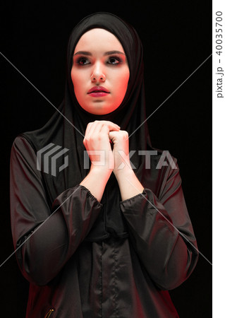 Portrait of beautiful serious young muslim woman wearing black hijab with hands near her face as 40035700