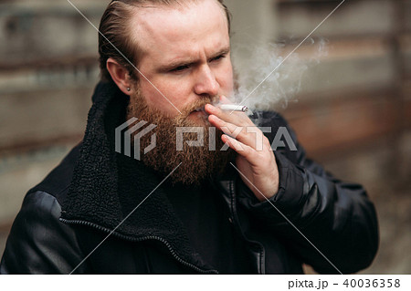 A bearded man in a black leather jacket stands outside and poses for a photographer. Smokes sigaret 40036358