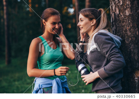 Two female runners relaxing after workout standing near the tree talking in park 40330665