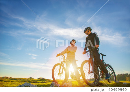 Cyclist couple with mountain bikes standing on the hill under the evening sky and enjoying bright Cyclist couple with mountain bikes standing on the hill under the evening sky and enjoying bright 40499940