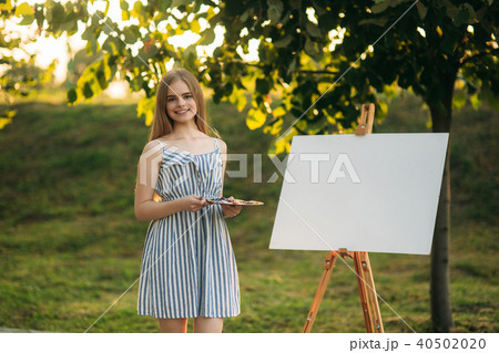 Beautiful girl standing in the park and draws a picture using a palette with paints and a spatula. Beautiful girl standing in the park and draws a picture using a palette with paints and a spatula. 40502020