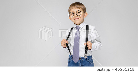 A charming boy in a white shirt, suspenders, a tie and light jeans stands on a gray background. the 40561319