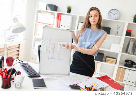 A beautiful young girl in an office stands near a table and explains on a magnetic blackboard a A beautiful young girl in an office stands near a table and explains on a magnetic blackboard a 40564973