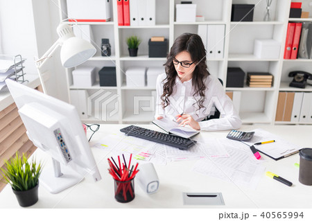 A young girl is working at the computer in the office and holds a pen and notebook in her hand. A young girl is working at the computer in the office and holds a pen and notebook in her hand. 40565994