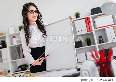 Beautiful young girl stands near an office desk and points with a pen on an empty board. 40566384