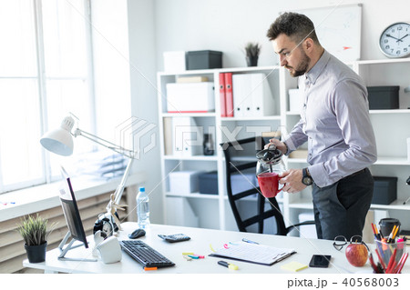 A young man in the office is standing near the table and pouring coffee into a cup. 40568003