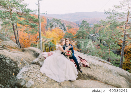 Horizontal view of the happy newlywed couple sitting on the knitted plaid on the rock. 40586241