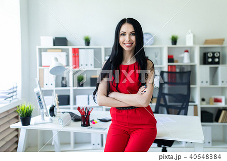 Beautiful young girl in a red suit is standing in the office, leaning on the table. Beautiful young girl in a red suit is standing in the office, leaning on the table. 40648384