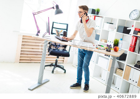 A young man in glasses stands near a computer desk and is talking on the phone. Before him lies a 40672889