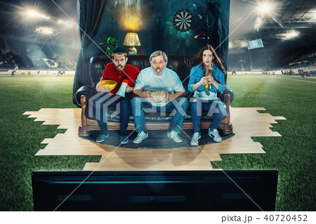 Soccer football fans sitting on the sofa and watching TV in the middle of a football field. 40720452