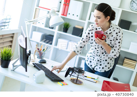 A young girl is standing in the office near the table, holding a red mug in her hand and typing on A young girl is standing in the office near the table, holding a red mug in her hand and typing on 40730840