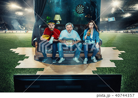 Soccer football fans sitting on the sofa and watching TV in the middle of a football field. 40744357