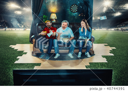 Soccer football fans sitting on the sofa and watching TV in the middle of a football field. Soccer football fans sitting on the sofa and watching TV in the middle of a football field. 40744391