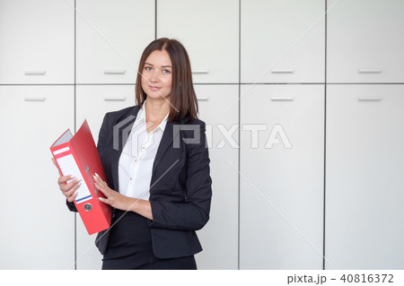 Beautiful business woman stands with a red folder in hands in a office Beautiful business woman stands with a red folder in hands in a office 40816372