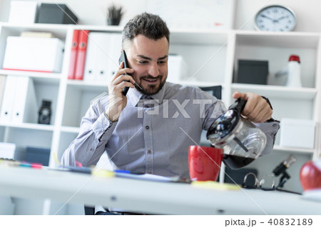 A young man in the office sits at a table, talking on the phone and pouring coffee into a cup. 40832189