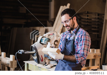 Handsome carpenter in protective glasses is looking at camera and smiling while standing near his 40899511