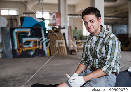 Handsome carpenter in protective glasses is looking at camera and smiling while standing near his 40900070