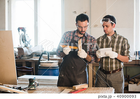 Happy male carpenter showing something to coworker at his notebook papers in workshop Happy male carpenter showing something to coworker at his notebook papers in workshop 40900093