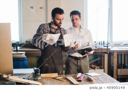 Happy male carpenter showing something to coworker at his notebook papers in workshop Happy male carpenter showing something to coworker at his notebook papers in workshop 40900149