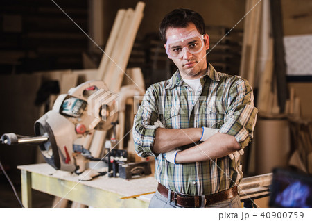 Handsome carpenter in protective glasses is looking at camera and smiling while standing near his 40900759