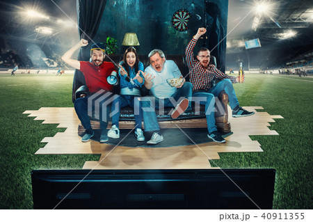 Soccer football fans sitting on the sofa and watching TV in the middle of a football field. 40911355