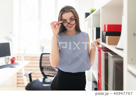 Beautiful young girl in an office stands near a shelf with documents and holds her hand glasses. Beautiful young girl in an office stands near a shelf with documents and holds her hand glasses. 40947707