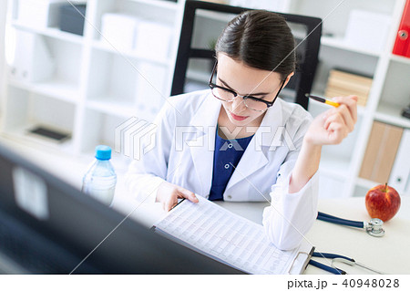 A beautiful young girl in a white robe is sitting at a computer desk with documents and a pen in her 40948028