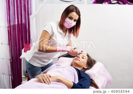 Cosmetologist is looking in the camera while giving facial treatment with a syringe to a client of a Cosmetologist is looking in the camera while giving facial treatment with a syringe to a client of a 40958594