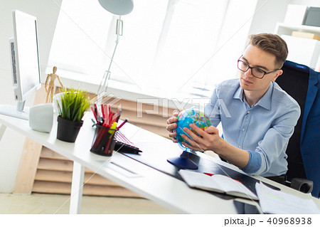 A young man sits in the office at a computer desk and holds a globe in his hands. 40968938