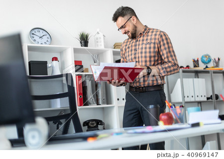 A man in the office stands near the shelf and scrolls through the folder with the documents. A man in the office stands near the shelf and scrolls through the folder with the documents. 40969147
