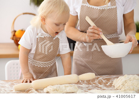 Little girl and her blonde mom in beige aprons  playing and laughing while kneading the dough in th 41005617