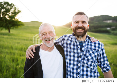 A portrait of an adult hipster son with senior father in nature at sunset, arms around each other. 41076643
