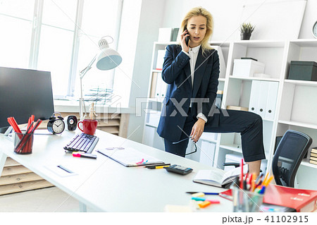 A young girl stands near a table in the office, putting her foot on a chair and talking on the phone 41102915