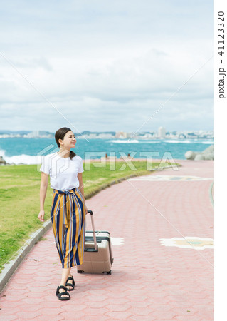 スーツケースを転がしながら海沿いの道を歩く女性の写真素材