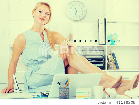young cheerful adult girl in blue dress sitting on table in home office young cheerful adult girl in blue dress sitting on table in home office 41138509