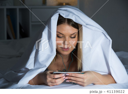 Female under blanket with mobile phone smiling on bed Female under blanket with mobile phone smiling on bed 41139022