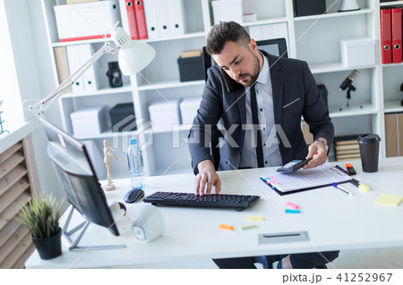 The man is sitting in the office at the desk, holding a calculator in his hand, supports the phone The man is sitting in the office at the desk, holding a calculator in his hand, supports the phone 41252967