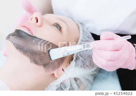 The cosmetologist in pink gloves with a brush applies a carbon mask for peeling on the face of a The cosmetologist in pink gloves with a brush applies a carbon mask for peeling on the face of a 41254022
