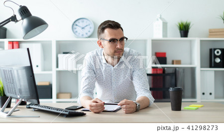 A young man in glasses stands near a table in the office and holds a pencil in his hands. A young man in glasses stands near a table in the office and holds a pencil in his hands. 41298273